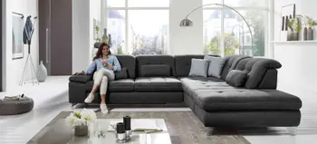 Frau sitzt auf dunkelgrauer Wohnlandschaft mit vielen Kissen, helles Wohnzimmer mit Panoramafenster, stilvolles Ambiente mit modernem Couchtisch.